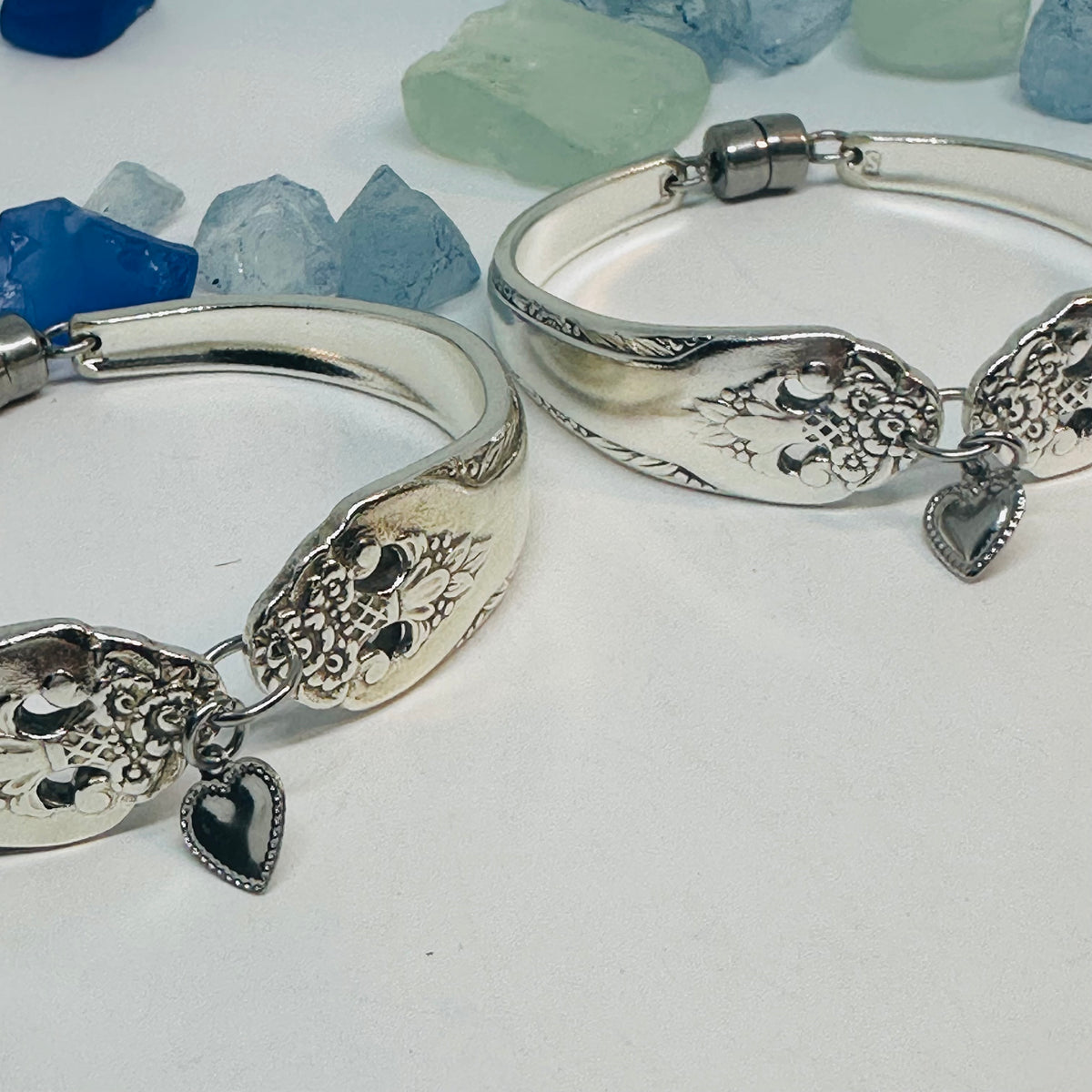 Two silver plated bangle bracelets featuring an engraved floral pattern and a dangling silver-tone heart charm, displayed on a white surface with blue and blue stones in the background.
