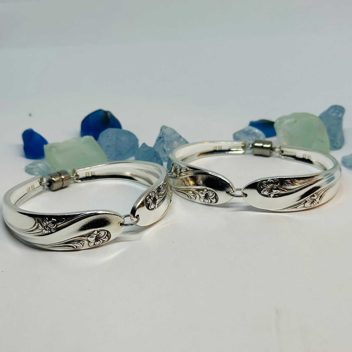 Two polished silver plated bracelets (Romance pattern) with engraved floral and leaf motifs, displayed on a light gray surface with decorative blue and green stones in the background.