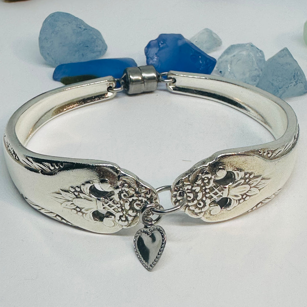 A silver plated bangle bracelet featuring an engraved floral pattern and a dangling silver-tone heart charm, displayed on a white surface with blue and blue stones in the background.
