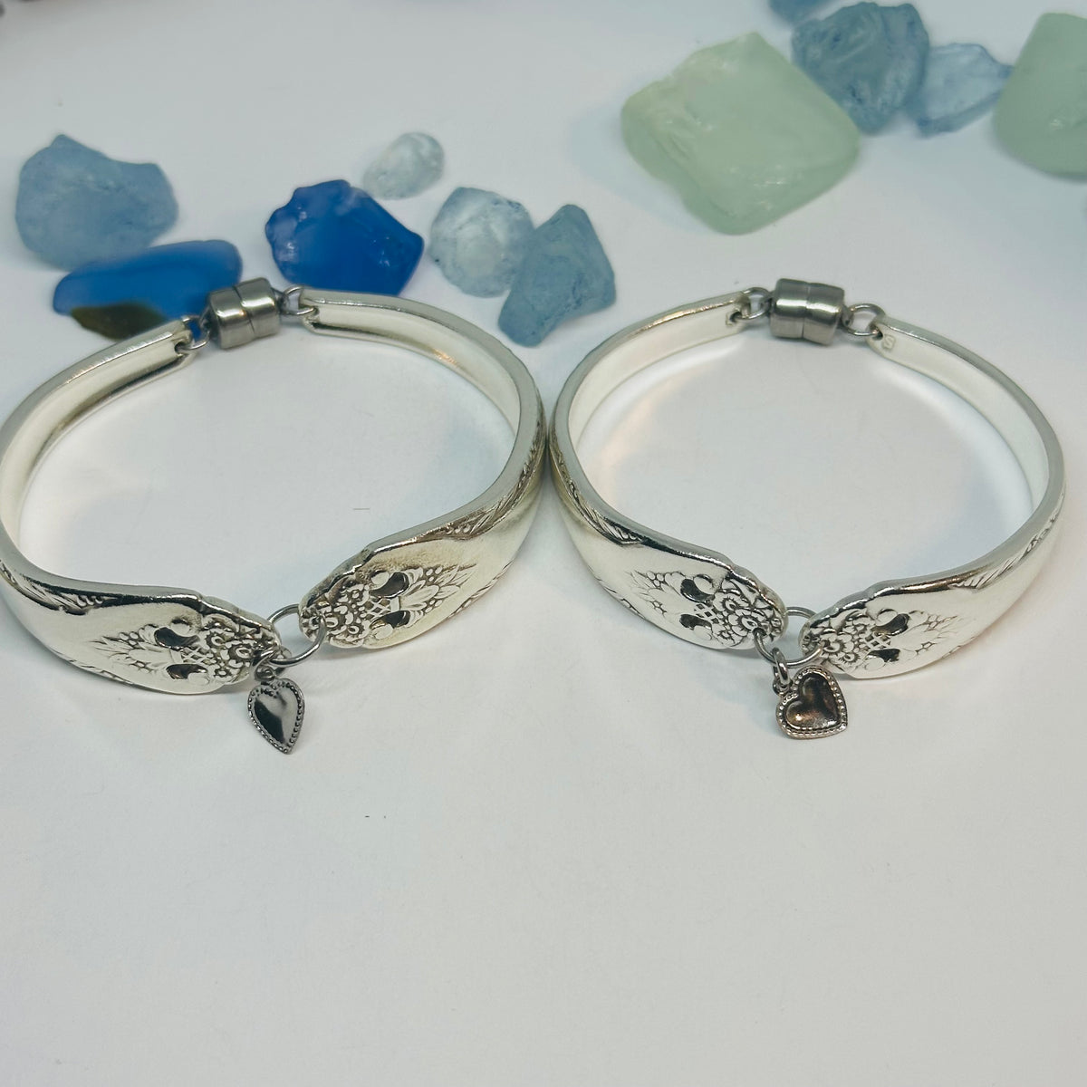 Two silver plated bangle bracelets featuring an engraved floral pattern and a dangling silver-tone heart charm, displayed on a white surface with blue and blue stones in the background.