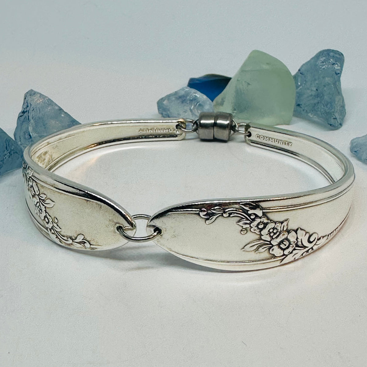 A silver plated spoon-link bangle bracelet "Queen Bess II" floral engravings, displayed on a white surface with decorative blue and green sea glass in the background. 