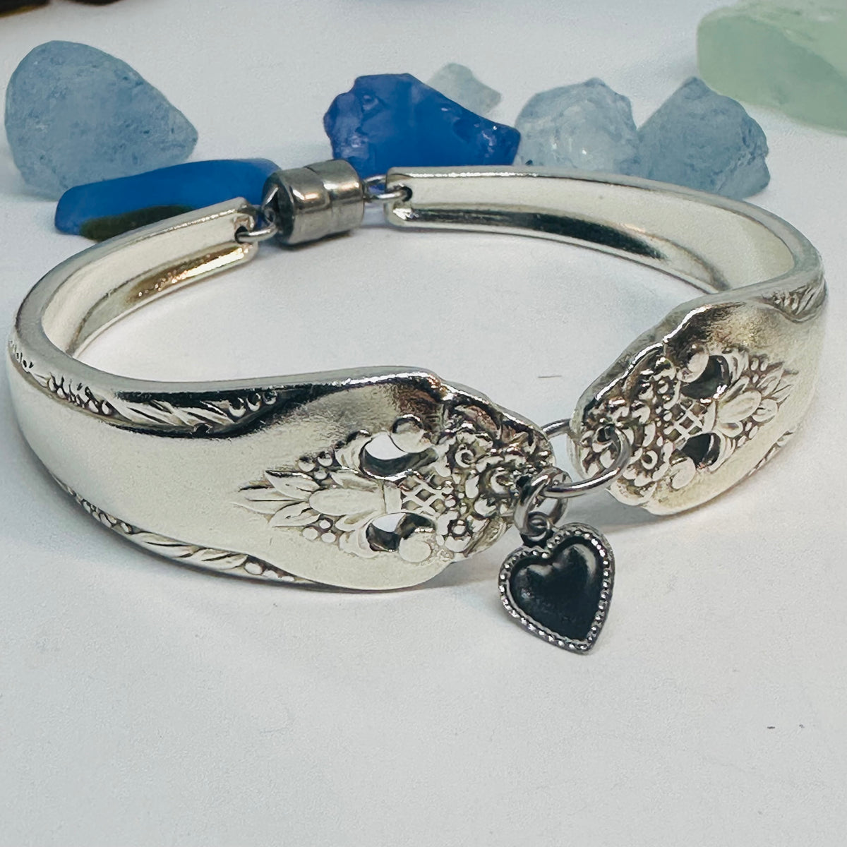 A silver plated bangle bracelet featuring an engraved floral pattern and a dangling silver-tone heart charm, displayed on a white surface with blue and blue stones in the background.