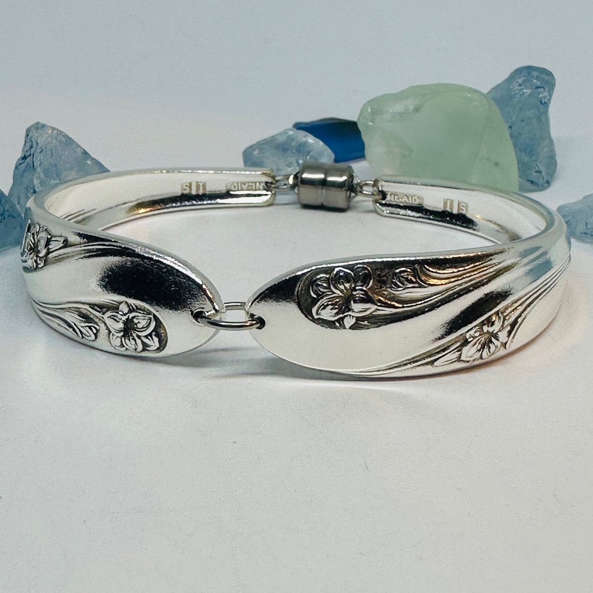 A polished silver plated bracelet (Romance pattern) with engraved floral and leaf motifs, displayed on a light gray surface with decorative blue and green stones in the background.