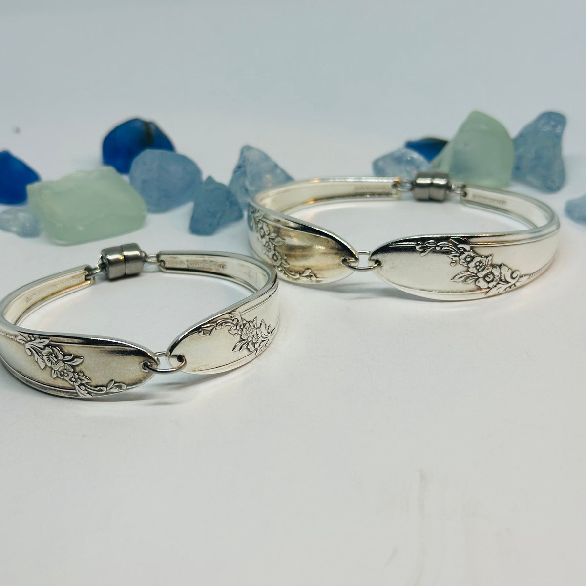 Two silver plated "Queen Bess II" bracelets with engraved floral details, displayed on a white surface with blue and green sea glass in the background.